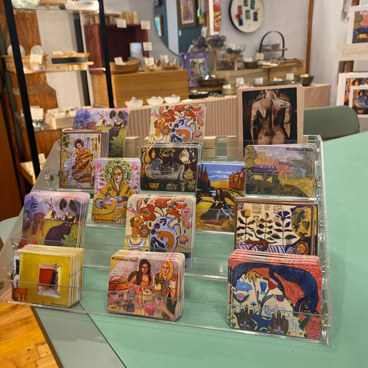 A clear acrylic card display stand holding multiple colorful art cards, photographed on a mint green table inside a shop with shelves and decor visible in the background.